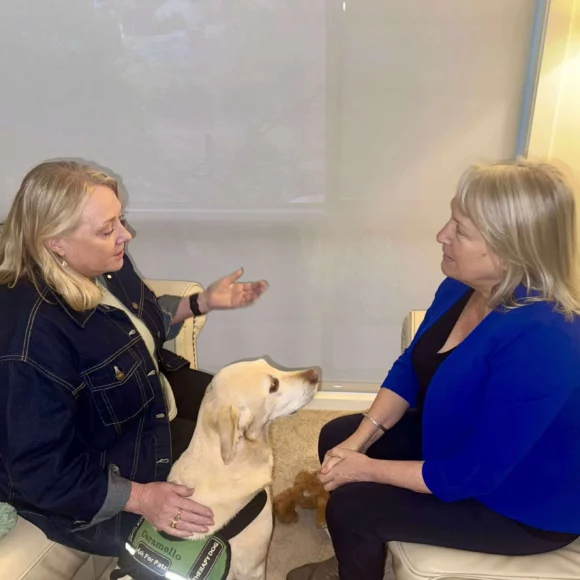 Two women sit facing each other, engaged in conversation. A yellow Labrador wearing a service dog vest sits between them, looking up attentively. The setting appears to be indoors with neutral-colored walls.