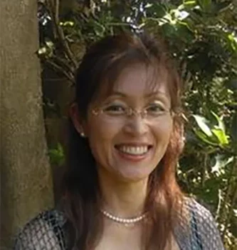 A woman with long brown hair and glasses smiles at the camera. She is wearing a pearl necklace and a textured top, standing outdoors with green foliage and trees in the background.