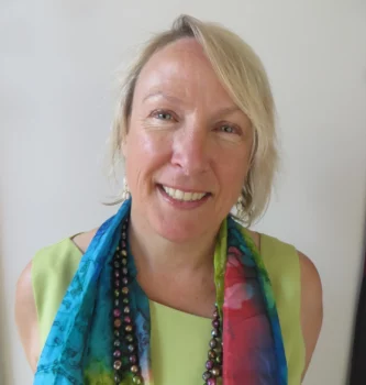 A smiling woman with short blonde hair wears a colorful scarf, a green top, and a beaded necklace, standing against a plain white background.