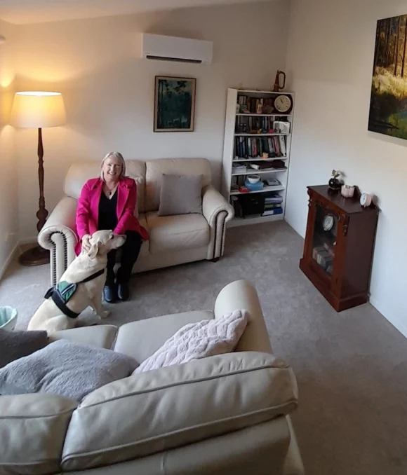 A woman in a pink jacket sits on a cream sofa in a cozy living room, smiling at a light-colored dog. There is another sofa, a bookshelf, a clock cabinet, a floor lamp, and a painting on the wall.