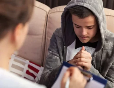 A teenage boy in a gray hoodie sits on a couch, looking down and clasping his hands, while an adult with a clipboard faces him, possibly during a counseling or therapy session.